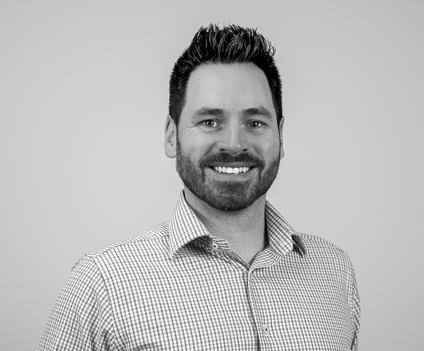 Black-and-white headshot of Jacob Ray, Partner, COO, and CCO at Colorado Wealth Group. He is smiling warmly, wearing a checkered dress shirt with the top button open. His hair is styled upward, and he has a neatly groomed beard. The background is plain and light-colored, giving a clean, professional look.