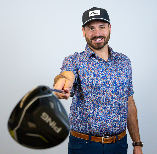 Jacob Ray, Partner, COO, and CCO at Colorado Wealth Group, posing playfully with a golf club. He is smiling and holding a Ping driver toward the camera. He’s wearing a patterned short-sleeve polo shirt, dark jeans, a leather belt, and a black-and-white ball cap. The background is a clean light gray, highlighting his casual and confident stance.