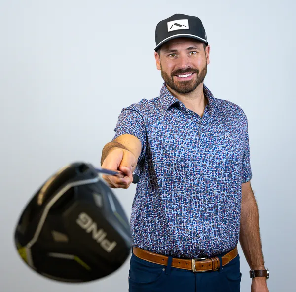 Jacob Ray, Partner, COO, and CCO at Colorado Wealth Group, posing playfully with a golf club. He is smiling and holding a Ping driver toward the camera. He’s wearing a patterned short-sleeve polo shirt, dark jeans, a leather belt, and a black-and-white ball cap. The background is a clean light gray, highlighting his casual and confident stance.