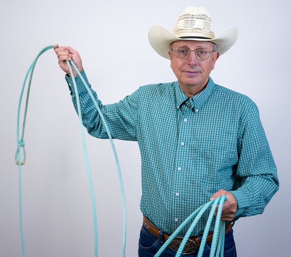 Bruce Larsen, Senior Wealth Advisor at Colorado Wealth Group, posing in a cowboy-themed outfit. He is wearing a white cowboy hat, teal patterned button-down shirt, jeans, and glasses. Bruce holds a coiled lasso in one hand and raises the loop in the other, smiling confidently in front of a plain light background.