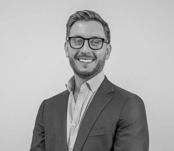 Black-and-white professional portrait of Matt Selihar, Wealth Advisor at Colorado Wealth Group. He is smiling broadly, wearing dark-rimmed glasses, a tailored suit jacket, and an open-collared dress shirt. His hair is neatly styled, and he stands confidently against a plain, light background.