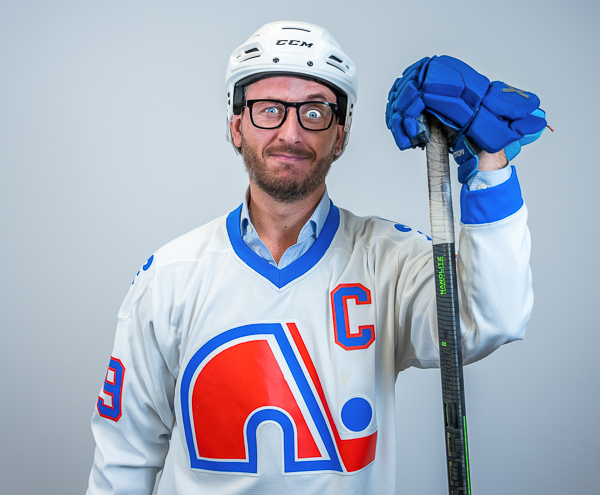 Matt Selihar, Wealth Advisor at Colorado Wealth Group, dressed in full hockey gear. He’s wearing a retro Quebec Nordiques jersey with a captain’s ‘C,’ blue hockey gloves, a white helmet, and black-rimmed glasses. He’s holding a hockey stick upright with one glove resting on top, standing confidently in front of a light gray background.