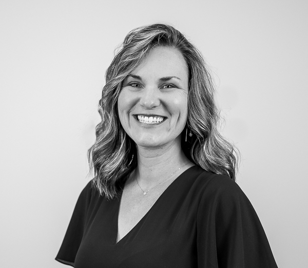 Black-and-white professional portrait of Emily Murtaugh, Director of Operations at Colorado Wealth Group. She is smiling warmly, wearing a dark V-neck blouse. Her medium-length wavy hair is styled loosely, and she stands against a plain, light background, creating a polished and approachable appearance.