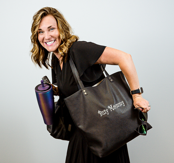Emily Murtaugh, Director of Operations at Colorado Wealth Group, smiling playfully while carrying a large black tote bag with 'Hot Mess' printed on it. She’s holding a rolled-up yoga mat and keys, dressed in a casual black outfit, and leaning forward in motion against a light gray background. Her expression is joyful and energetic.