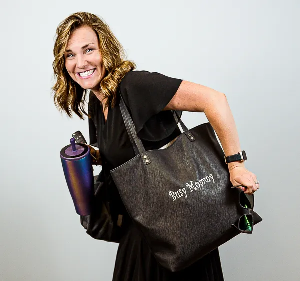 Emily Murtaugh, Director of Operations at Colorado Wealth Group, smiling playfully while carrying a large black tote bag with 'Hot Mess' printed on it. She’s holding a rolled-up yoga mat and keys, dressed in a casual black outfit, and leaning forward in motion against a light gray background. Her expression is joyful and energetic.