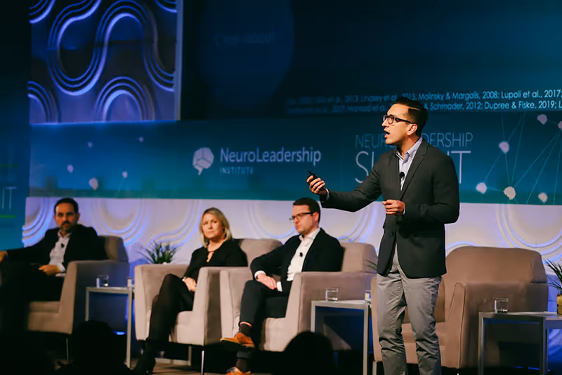 Speaker in a gray blazer presents on stage at NeuroLeadership Summit with three seated panelists behind.