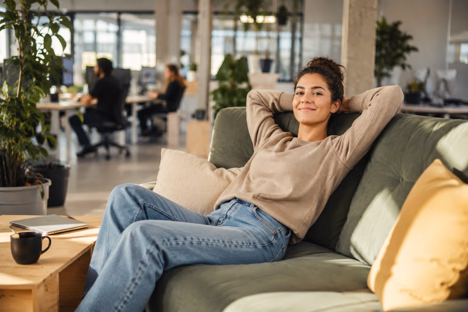 Eine junge Frau sitzt auf einer Couch in einem offenen Büro und lächelt zufrieden in die Kamera. Mit der epostbox entfällt der gesamte Platzbedarf für die Inhouse Produktionsinfrastruktur wie Drucker, Kuvertier- und Frankiermaschinen sowie der Lagerbedarf und schafft wieder Platz für Aufenthaltsqualität.