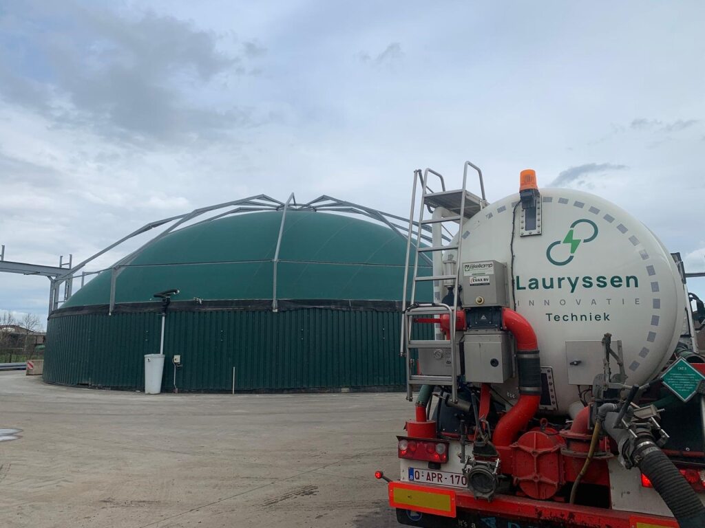 Een grote groene industriële koepel met metalen steunen staat onder een bewolkte hemel. Op de voorgrond staat een bedrijfswagen met slangen en ladders geparkeerd.