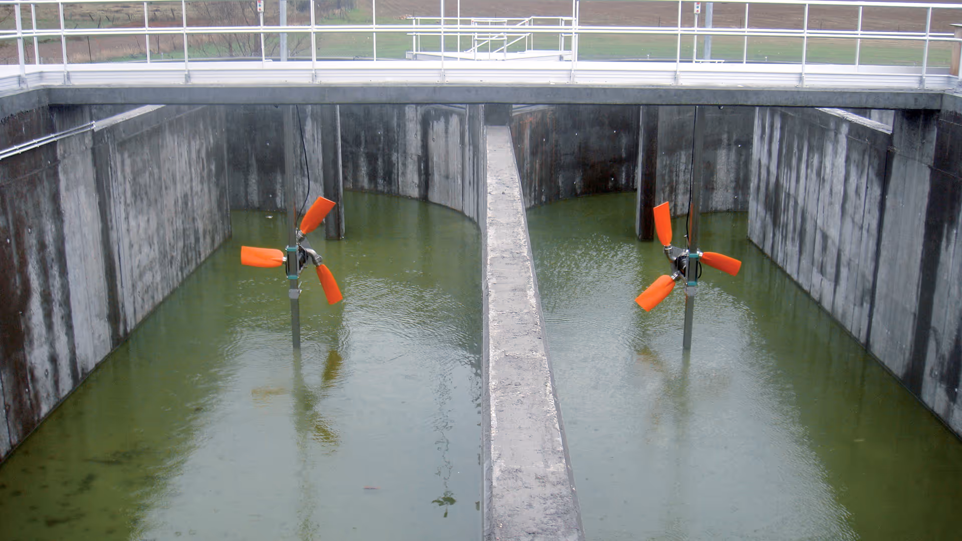 Een grote industriële waterzuiveringstank met groen algenrijk water, voorzien van twee rode, driebladige mixers. De betonnen wanden en structuur zijn zichtbaar.