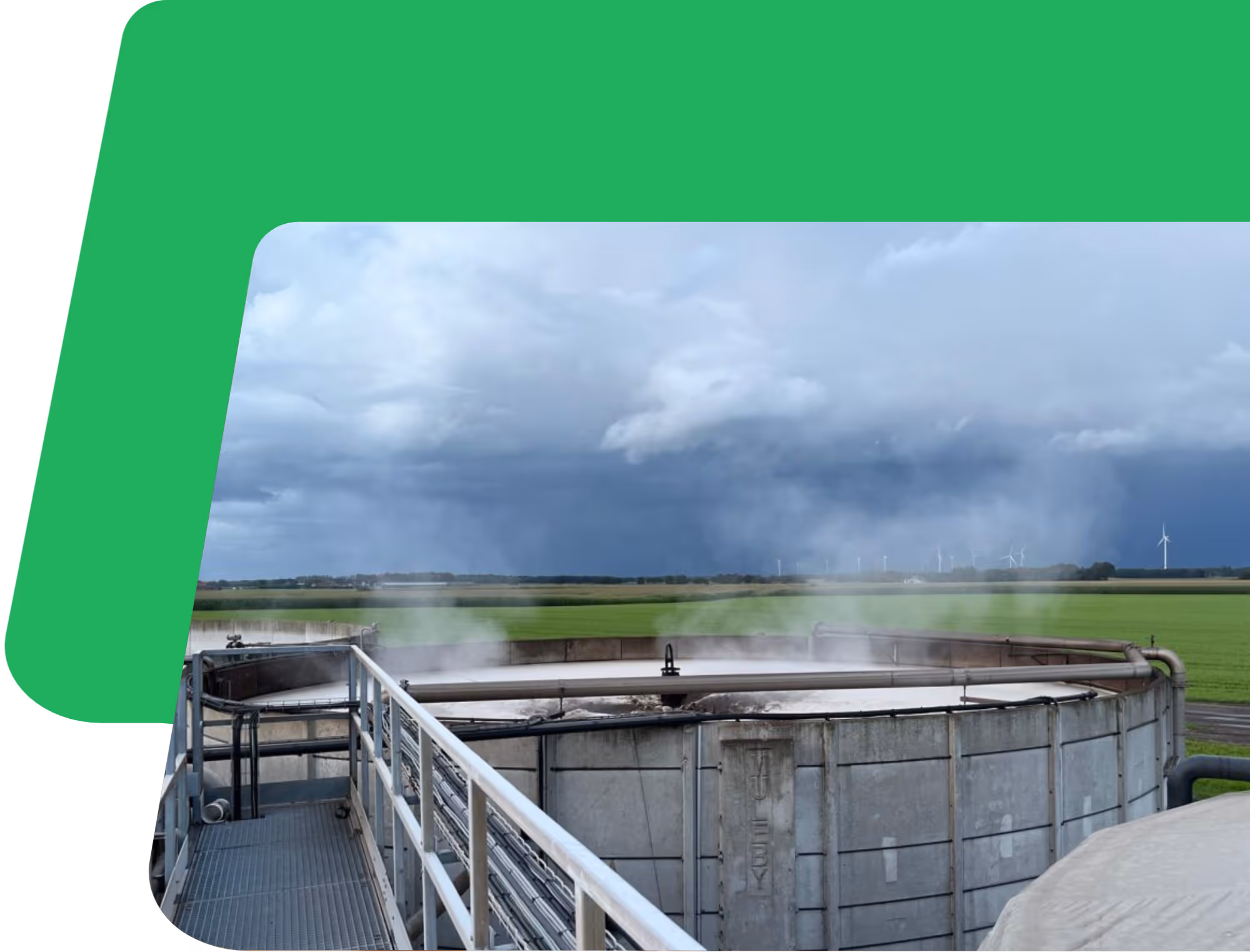 Grote industriële tank die stoom uitstoomt onder een bewolkte hemel, met windturbines en groene velden op de achtergrond. De scène voelt industrieel en toch sereen aan.