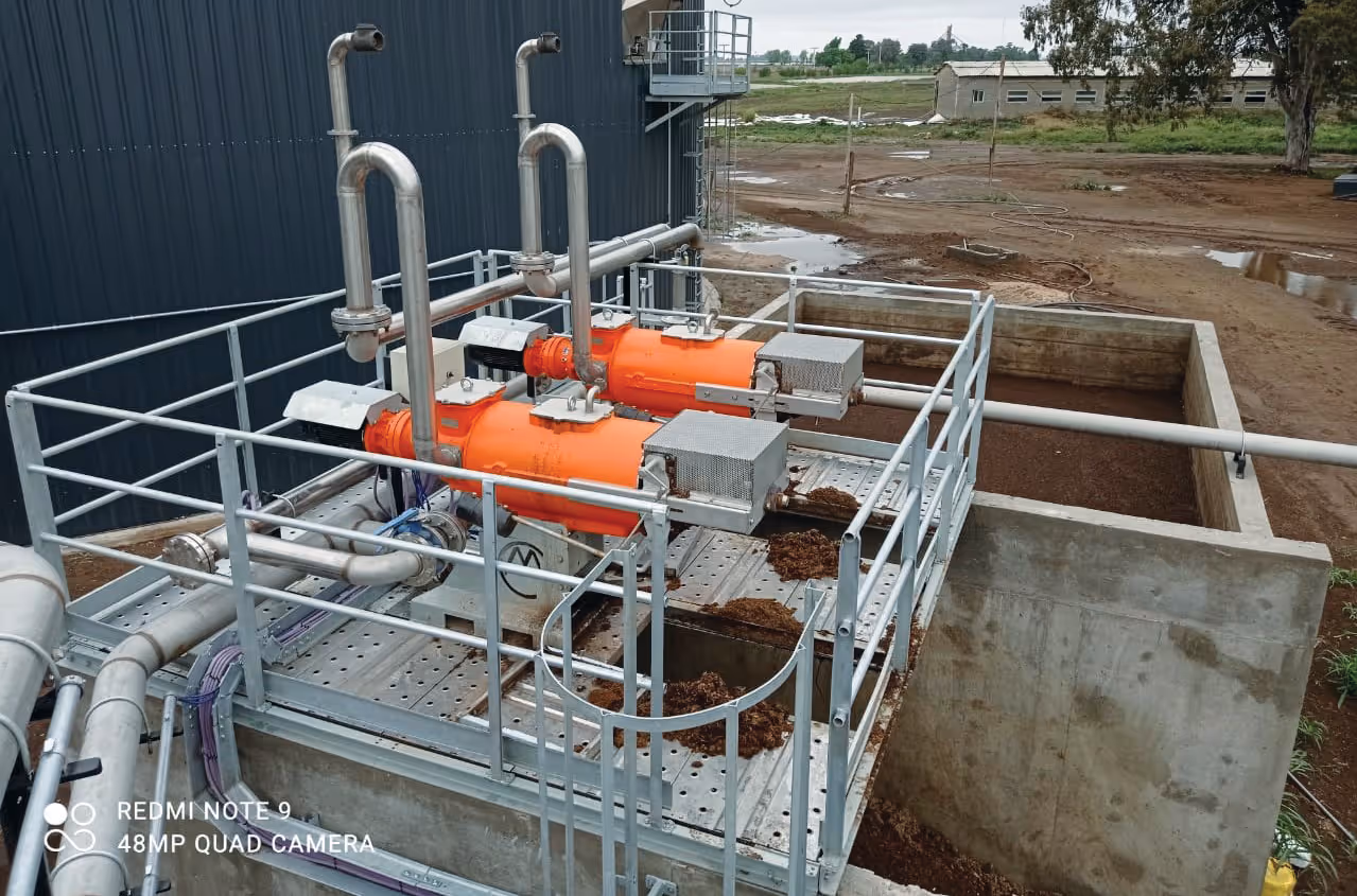 Industriële opstelling met grote oranje pompen en zilveren buizen op een platform, omgeven door hekwerk. Op de achtergrond is een modderige bouwplaats te zien.