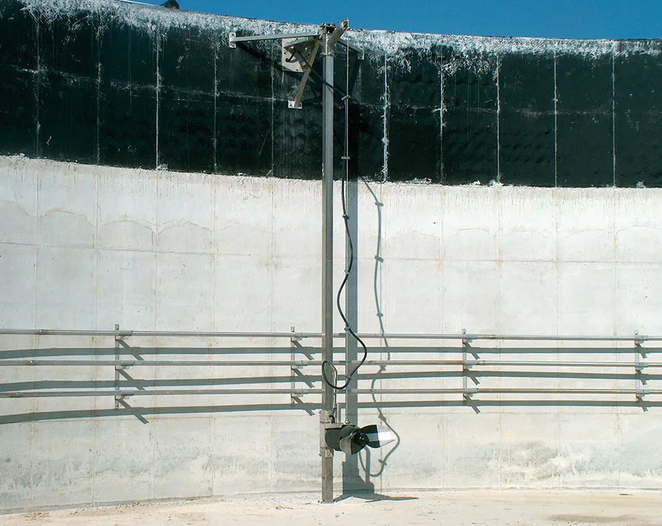 Stalen paalconstructie in een grote, lege betonnen tank met horizontale leuningen. De omgeving is industrieel onder een helderblauwe hemel.