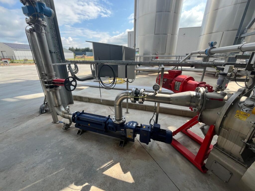 Industriële opstelling met metalen buizen, kleppen en machines onder een blauwe lucht. Grote tanks en een rode motor geven een efficiënte, drukke omgeving weer.