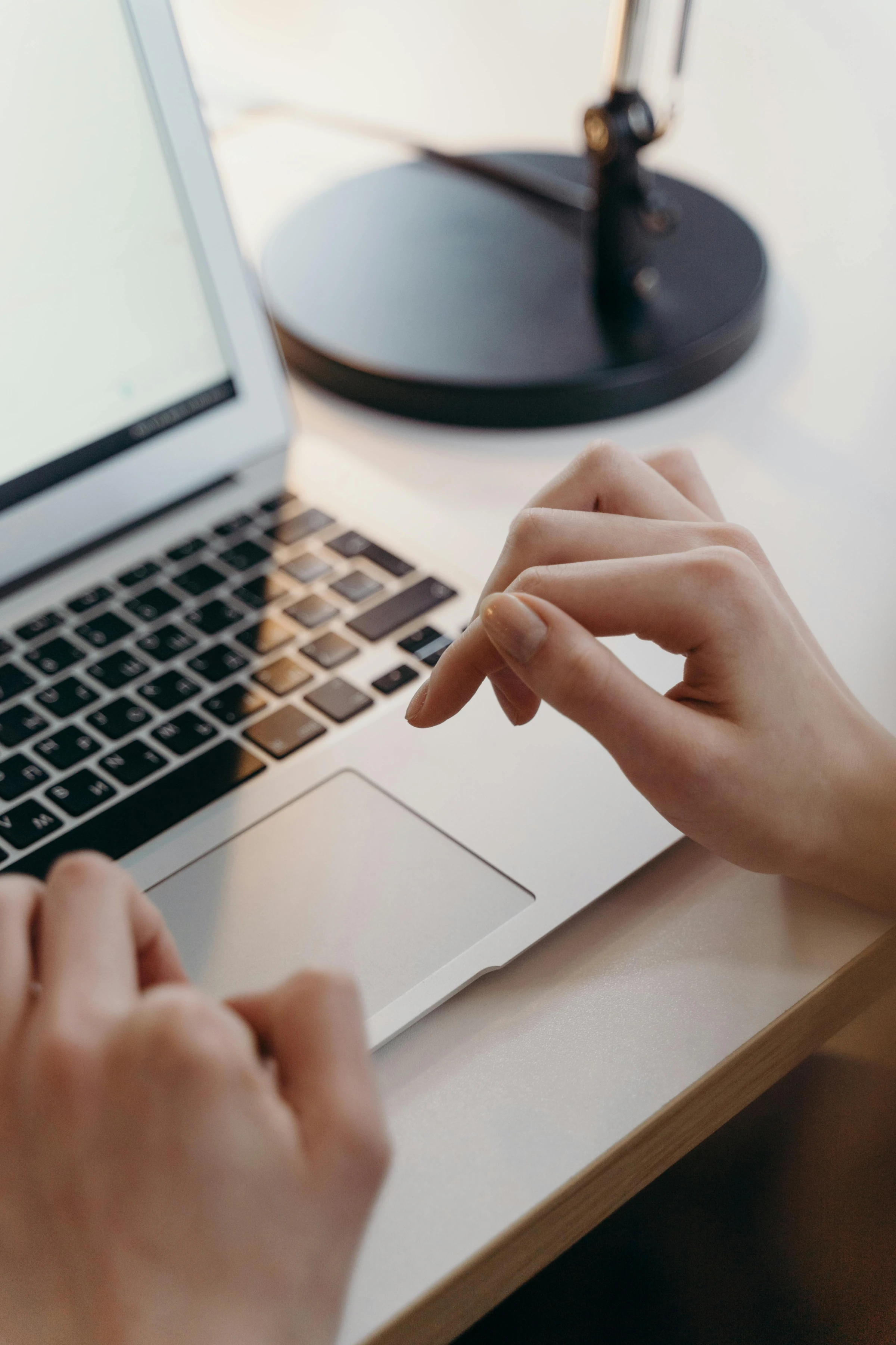 Close-up of hands typing on a laptop keyboard on a desk with a black lamp in the background precisely contracts