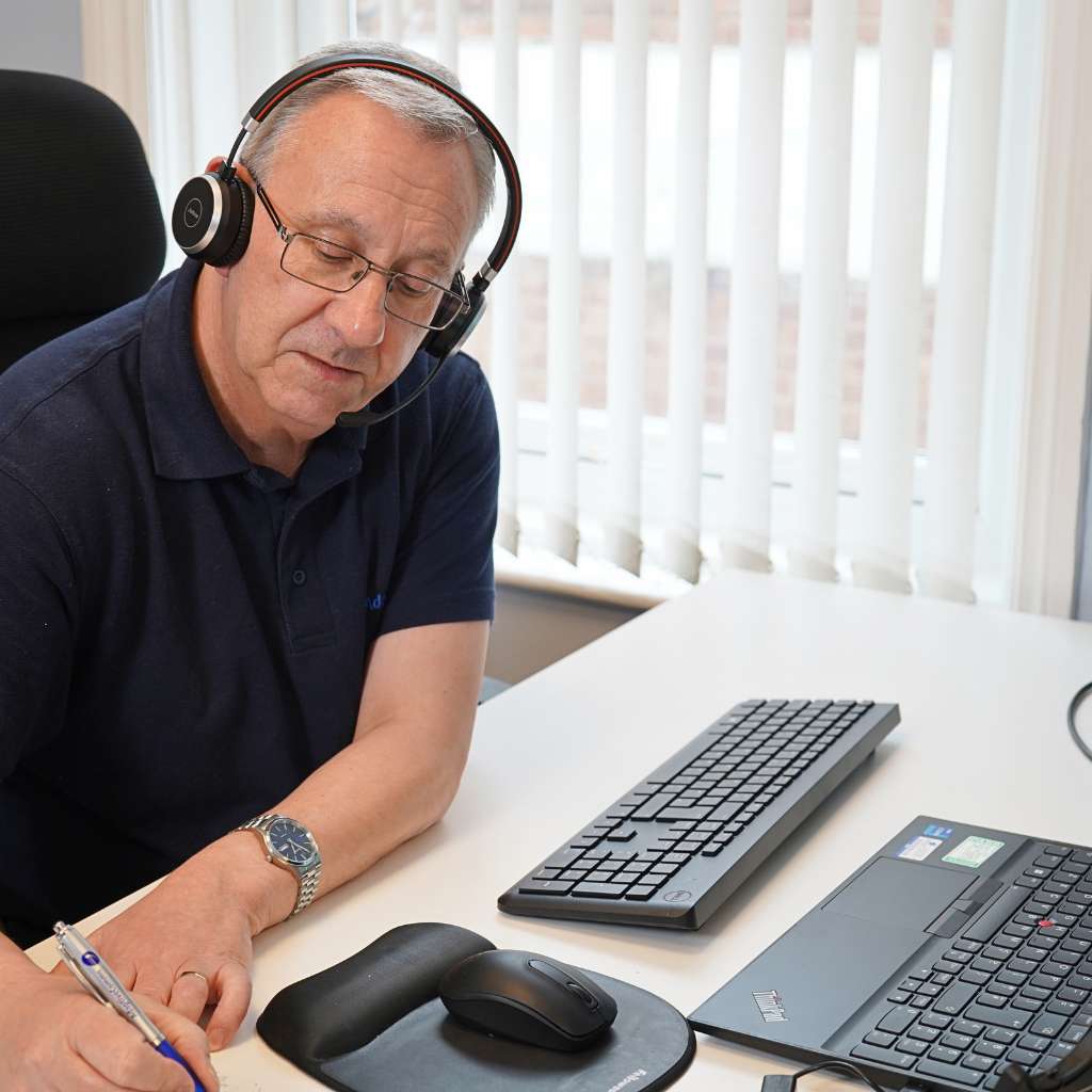 Man wearing headset writing notes at a desk with a laptop, keyboard, and mouse.