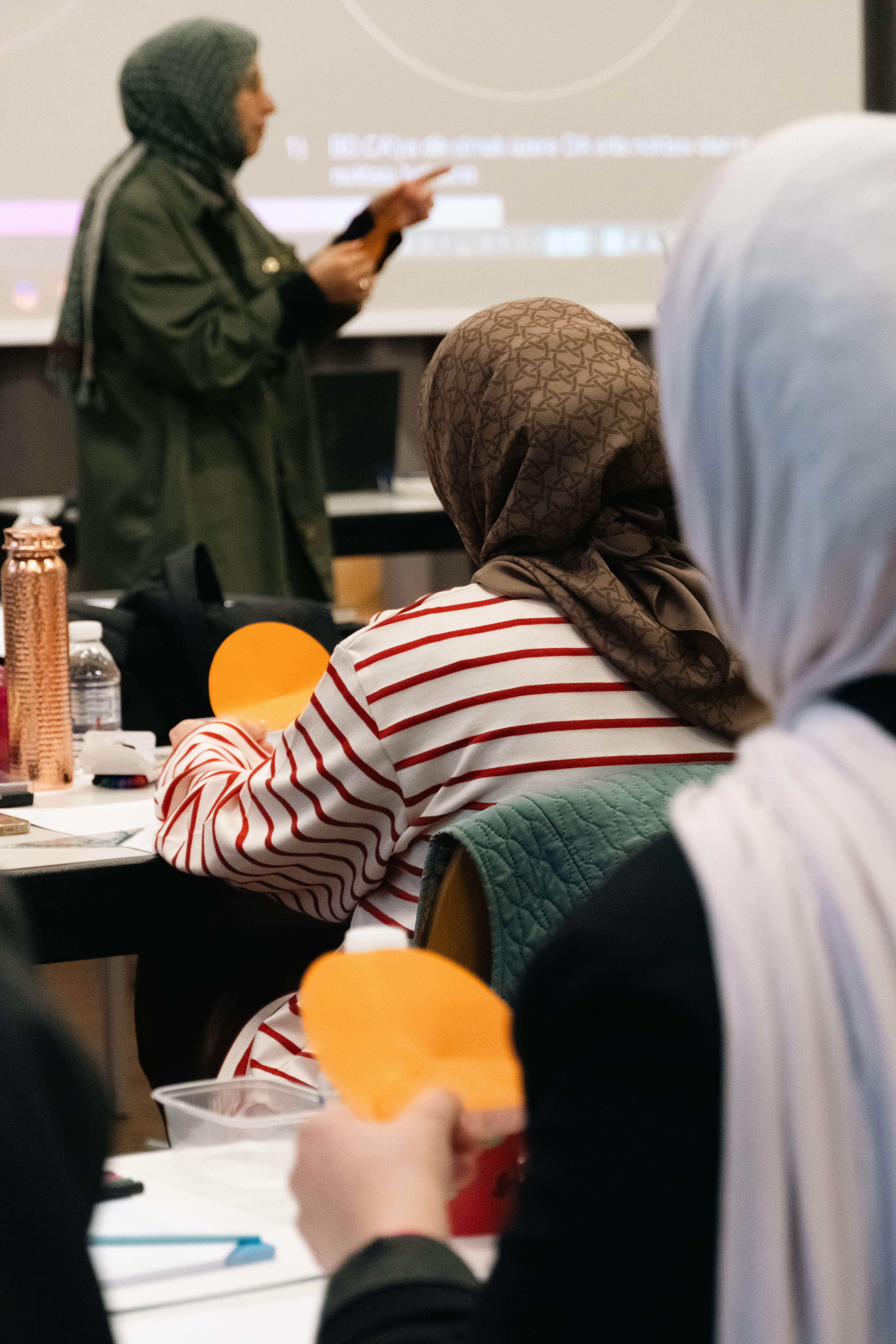 Students wearing hijabs attend a classroom session with a woman presenting in front of a projection screen.