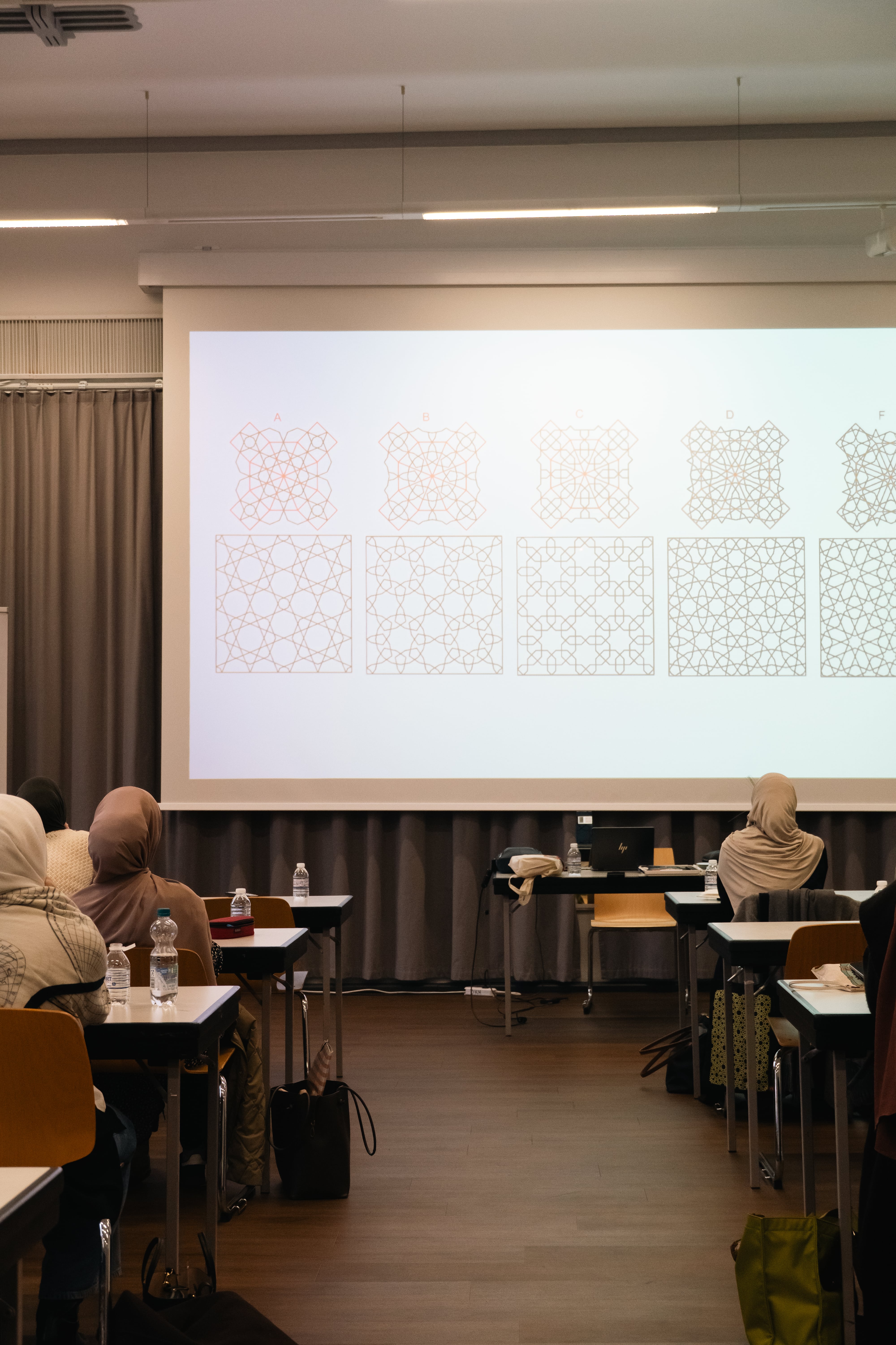 Classroom with students wearing headscarves facing a projector screen displaying six geometric pattern designs labeled A to F.