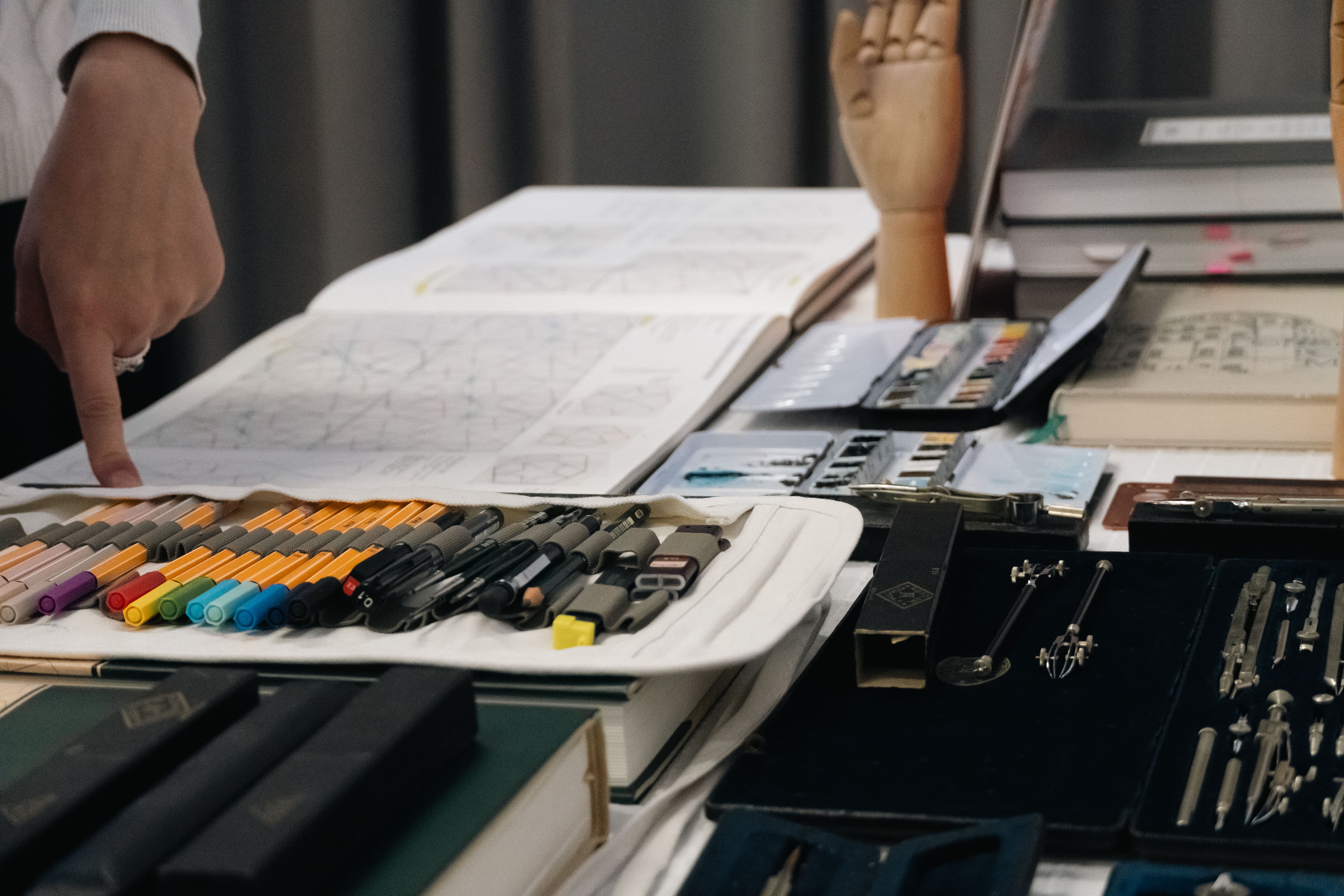 Close-up of drawing and drafting tools, colored pens, technical pens, compasses, and a hand pointing at a sketchbook with geometric drawings.