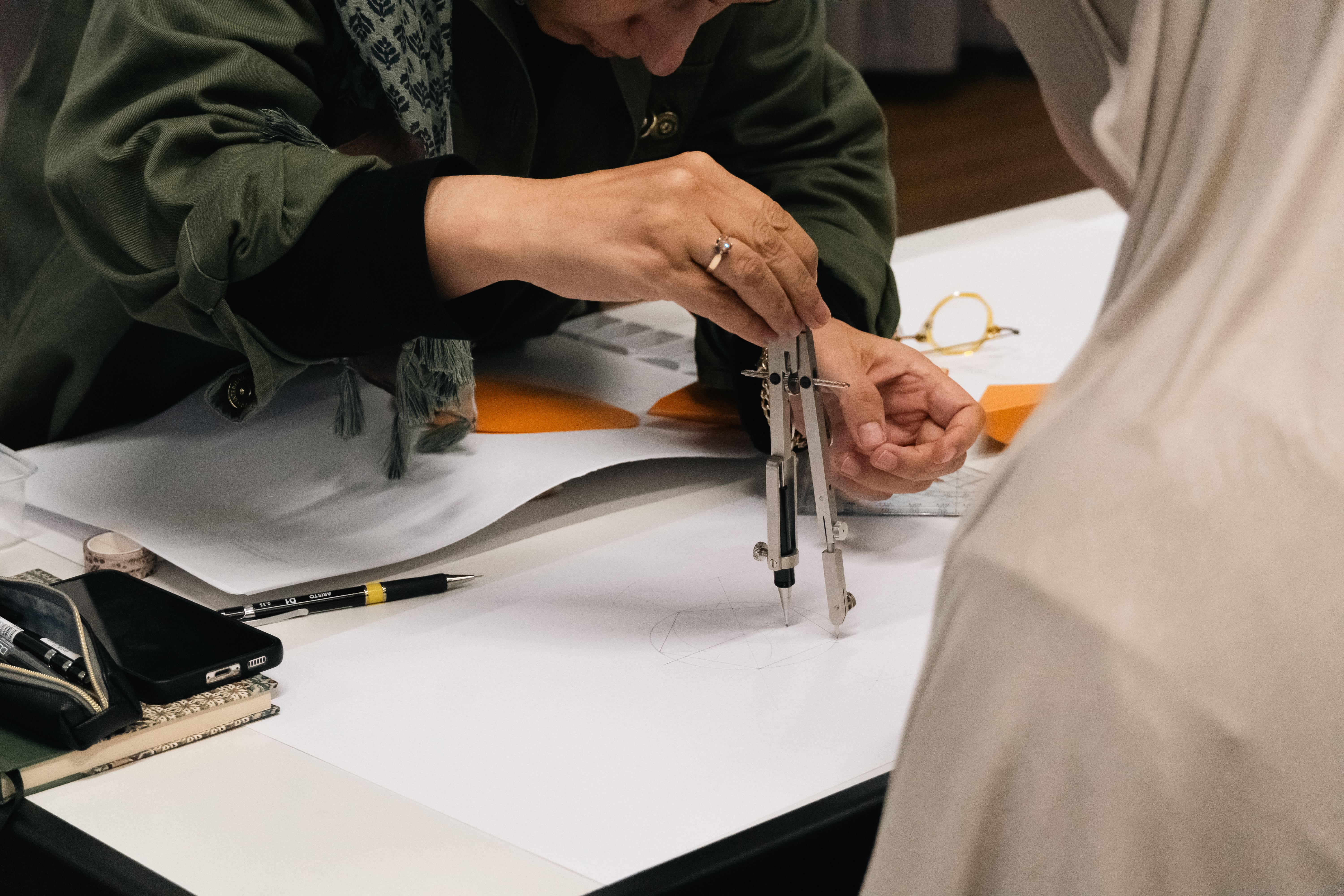 Person using a compass to draw on a white sheet of paper with another person partially visible nearby.