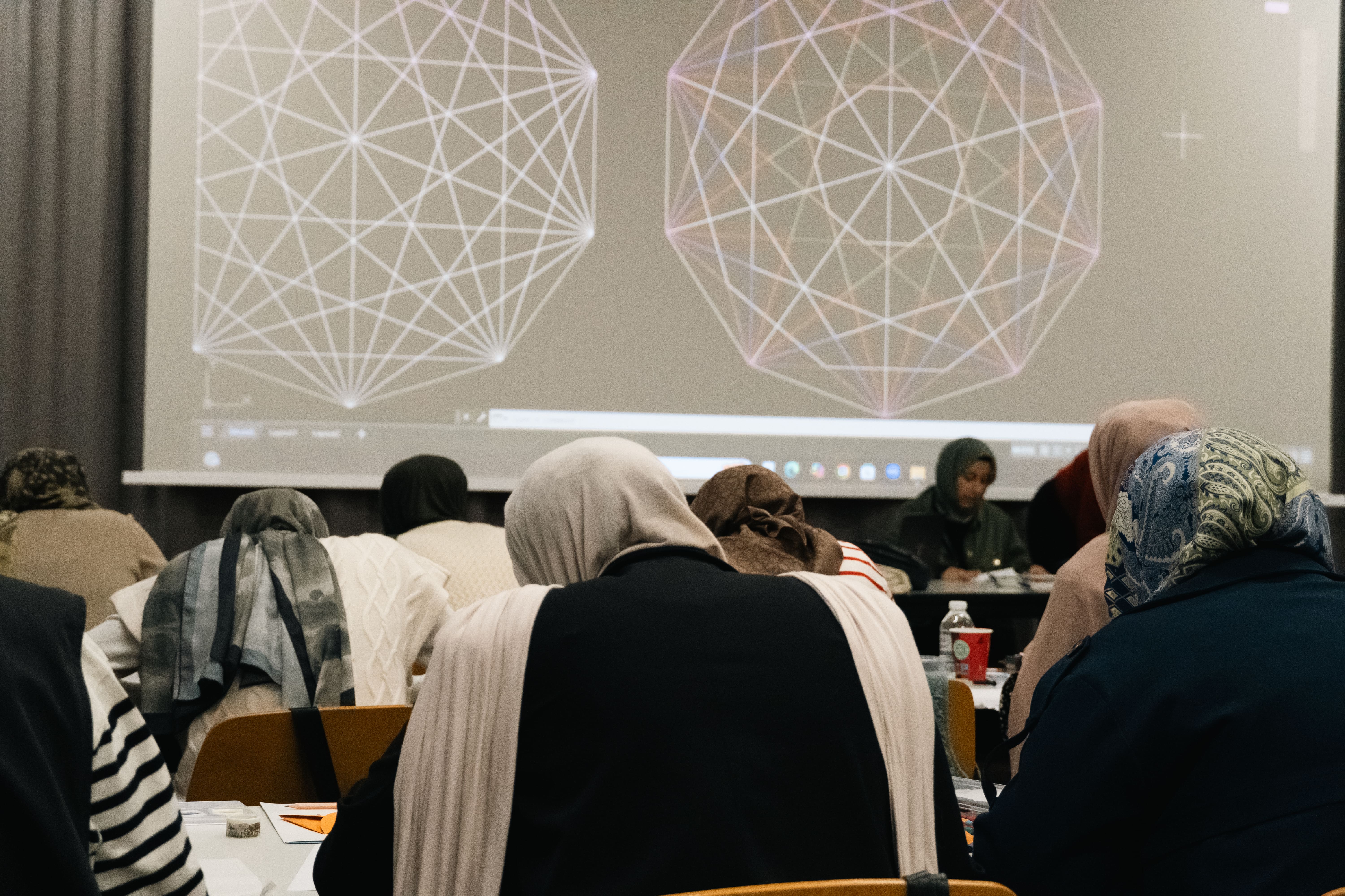 People wearing headscarves sitting in a classroom facing a screen displaying geometric wireframe shapes.