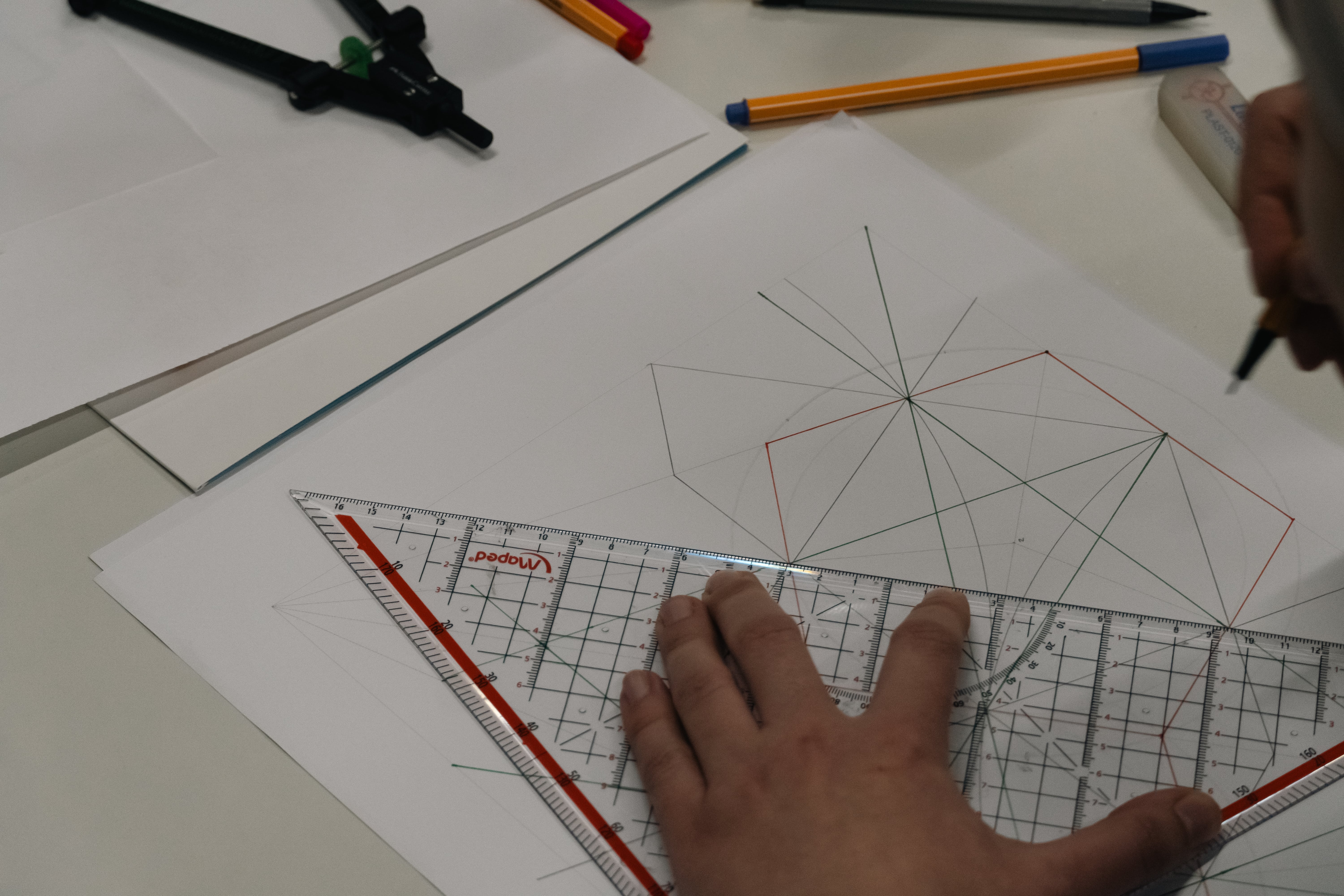 Hand holding a transparent set square ruler on a geometric drawing with colored lines on white paper.