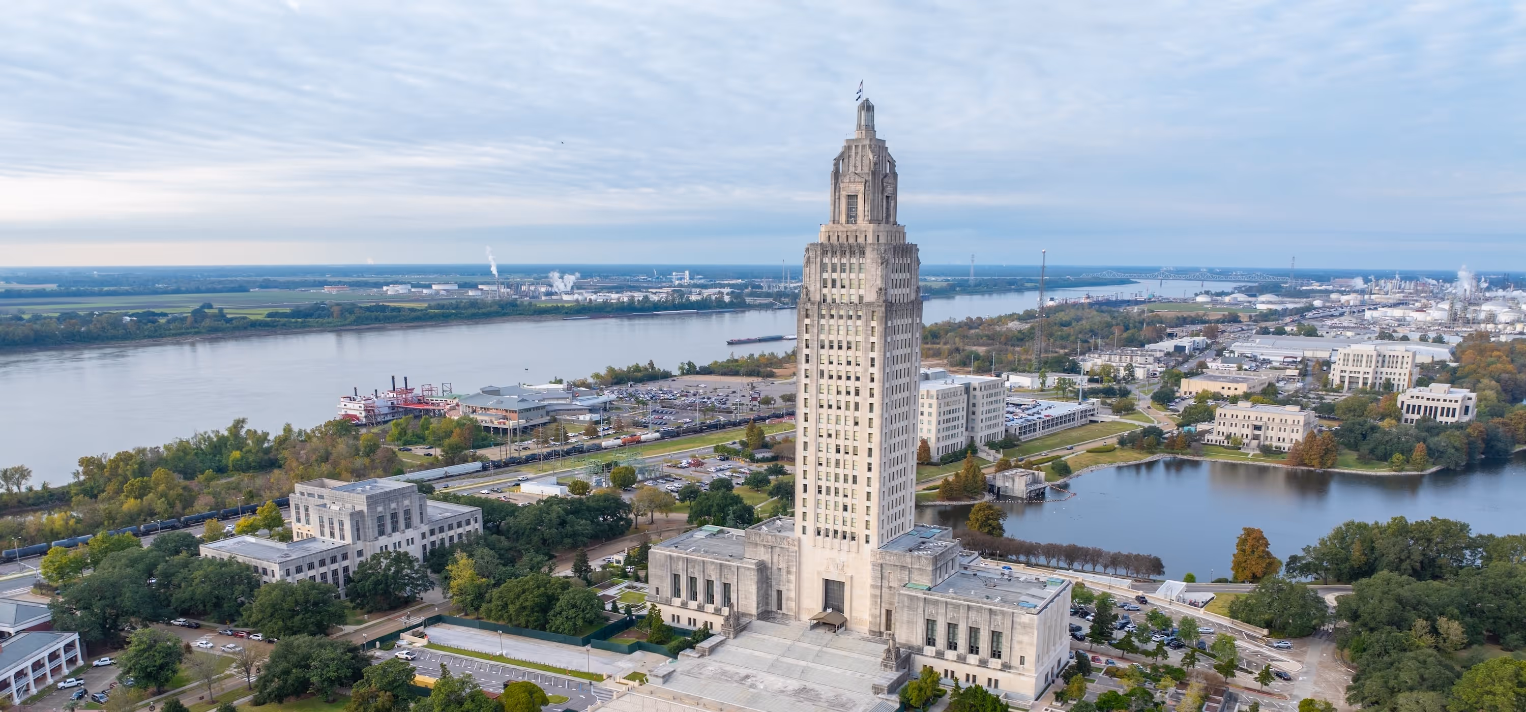 Aerial view of the Louisiana State Capitol building with a river and industrial area in the background under a cloudy sky.