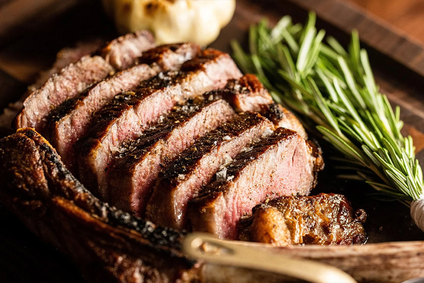 Sliced medium-rare steak on a wooden board with garlic bulb and fresh rosemary.