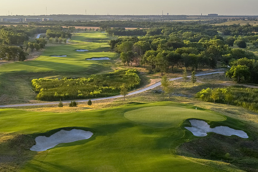 Scenic golf course with sand bunkers, green fairways, trees, and a distant horizon under a clear sky.
