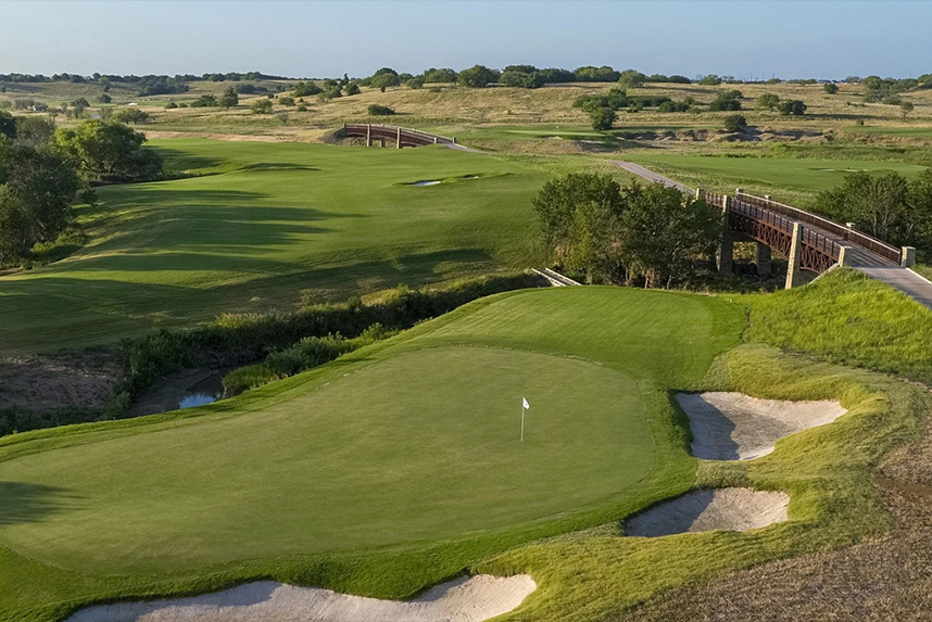 Golf course green with flagstick, sand bunkers, and a wooden bridge in a rolling landscape.