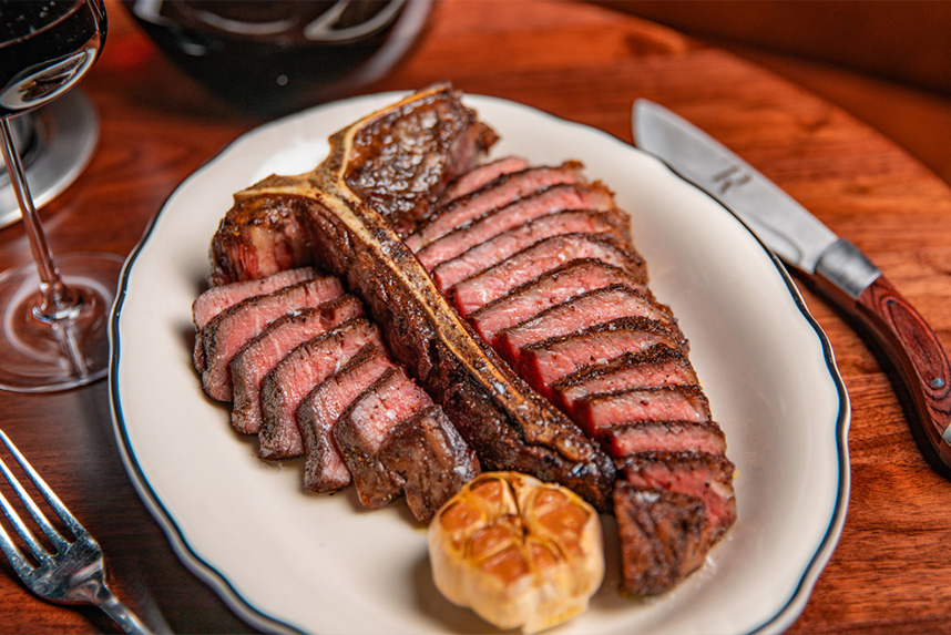 Sliced grilled T-bone steak on white plate with roasted garlic bulb, knife, fork, and wine glass.