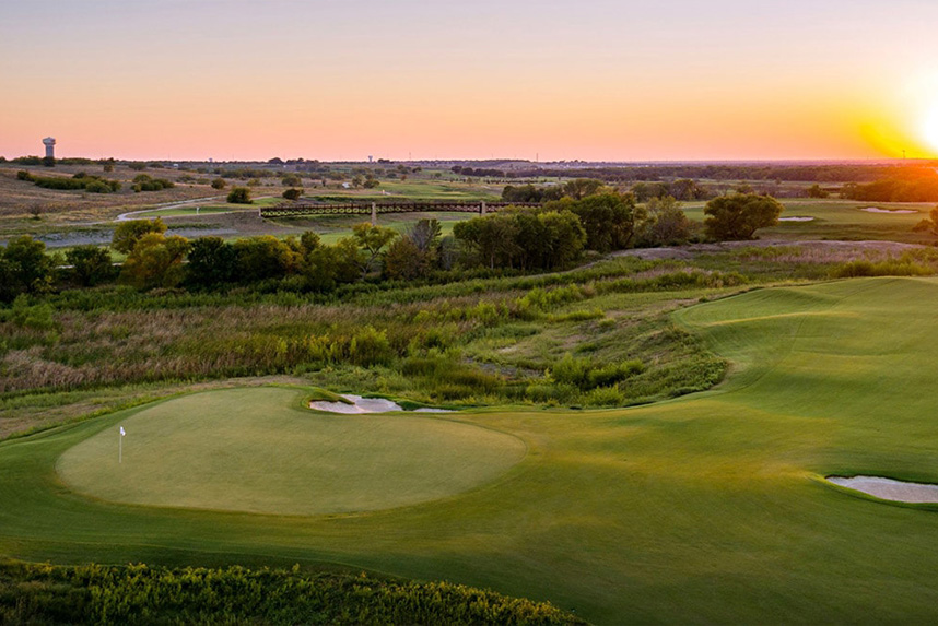 Sunset view of a golf course with greens, sand traps, trees, and a distant water tower.