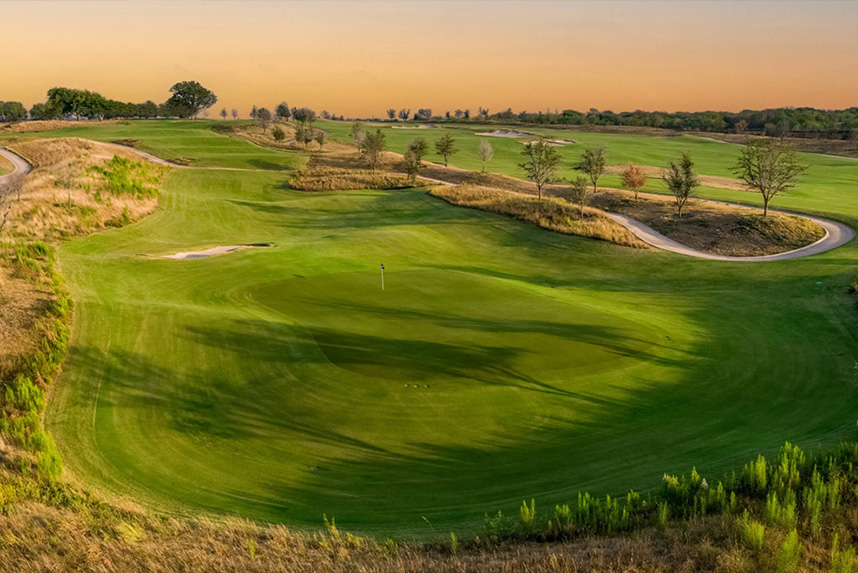 Wide view of a golf course green at sunset with a flagstick and trees lining a cart path.