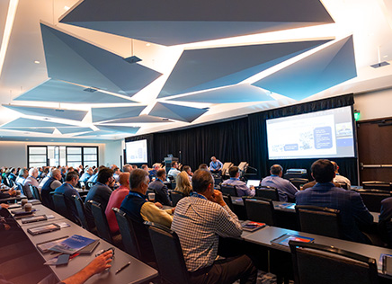 Audience seated in a modern conference room watching a presentation on two large screens.