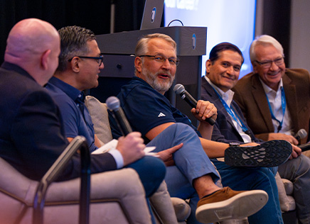 Five men sitting and talking on stage at a panel discussion, holding microphones.