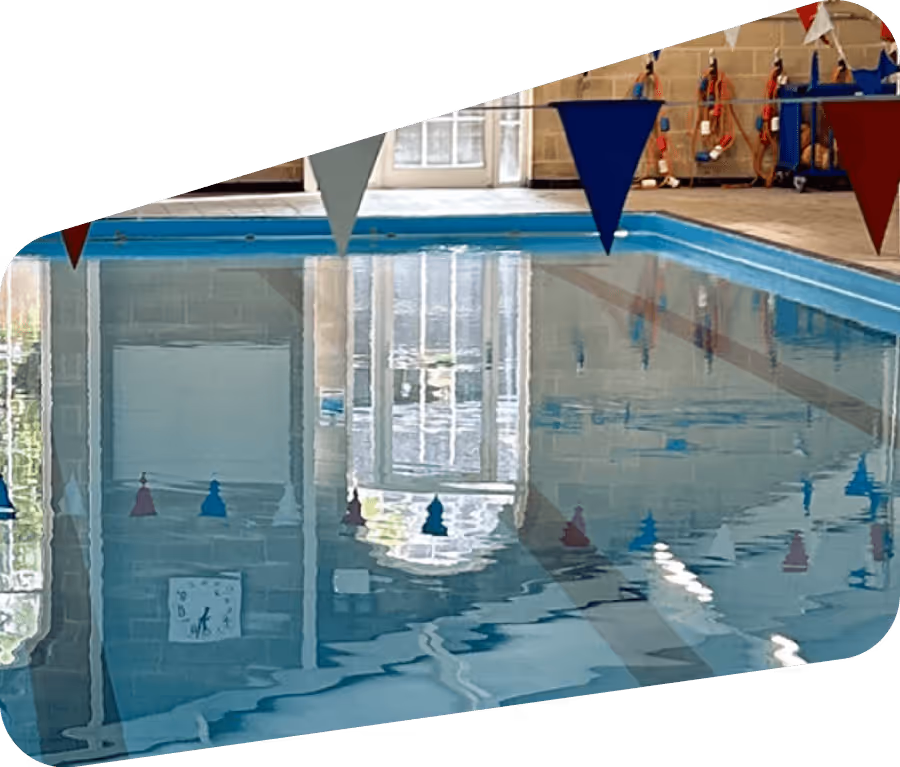 Indoor swimming pool with blue water, colorful triangular flags overhead, and reflections of windows and walls in the water.