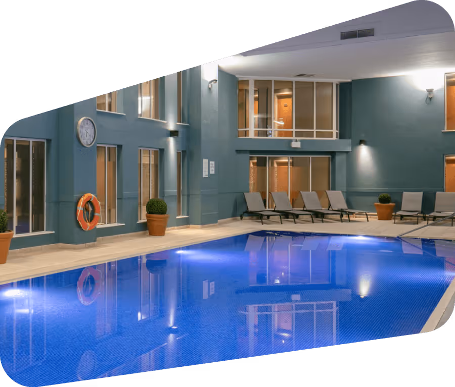 Indoor swimming pool with blue water, poolside lounge chairs, potted plants, and wall clock in a modern building.