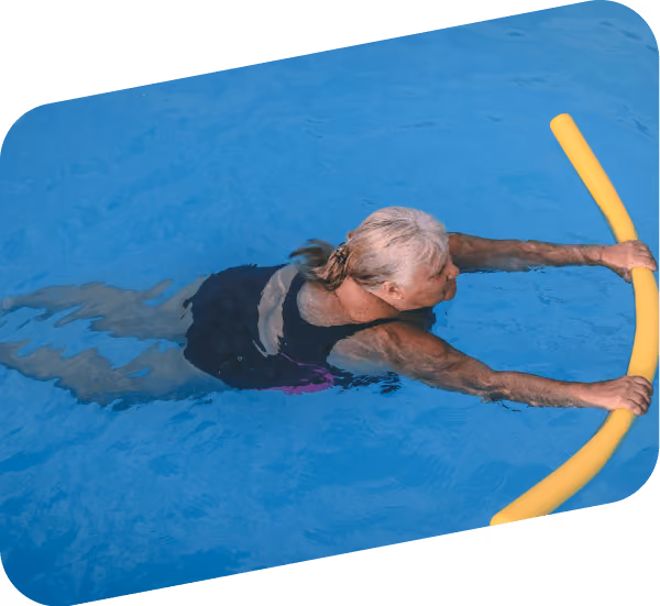 An older woman in a black swimsuit holds a yellow pool noodle while swimming in a clear blue pool.