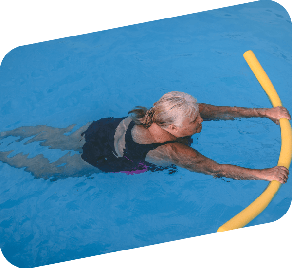 An older woman in a black swimsuit holds a yellow pool noodle while swimming in a clear blue pool.
