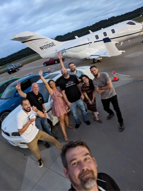 Selfie of Enthusiast Auto employees in front of an array of modern and vintage BMW's and a private jet