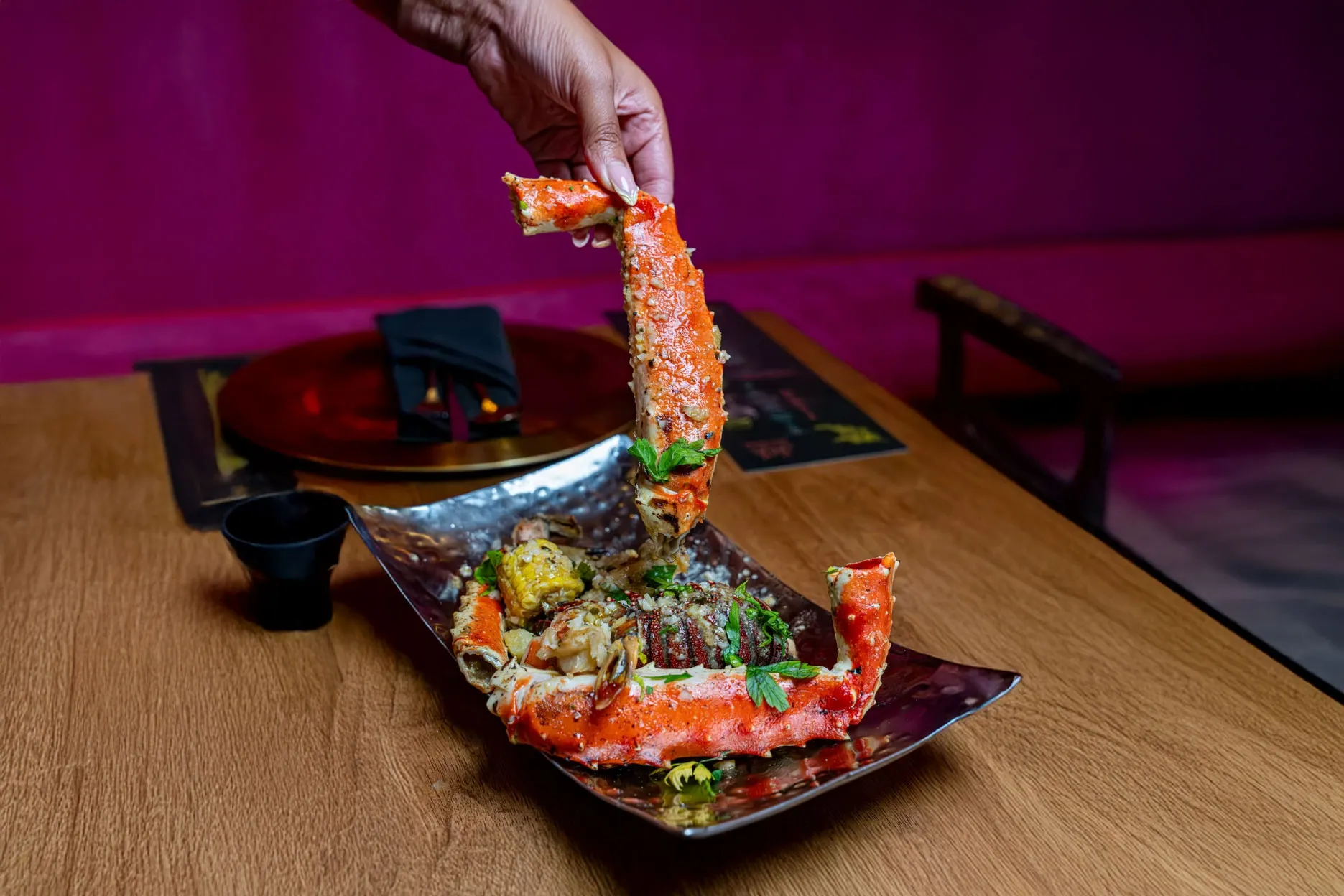 A hand lifts a large, vibrant red crab leg from a metal platter on a wooden table. The dish includes corn and herbs, against a deep magenta backdrop.