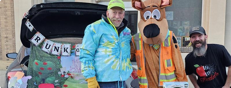 two people posing next to a person in a scooby doo costume in front of a car trunk filled with halloween treats