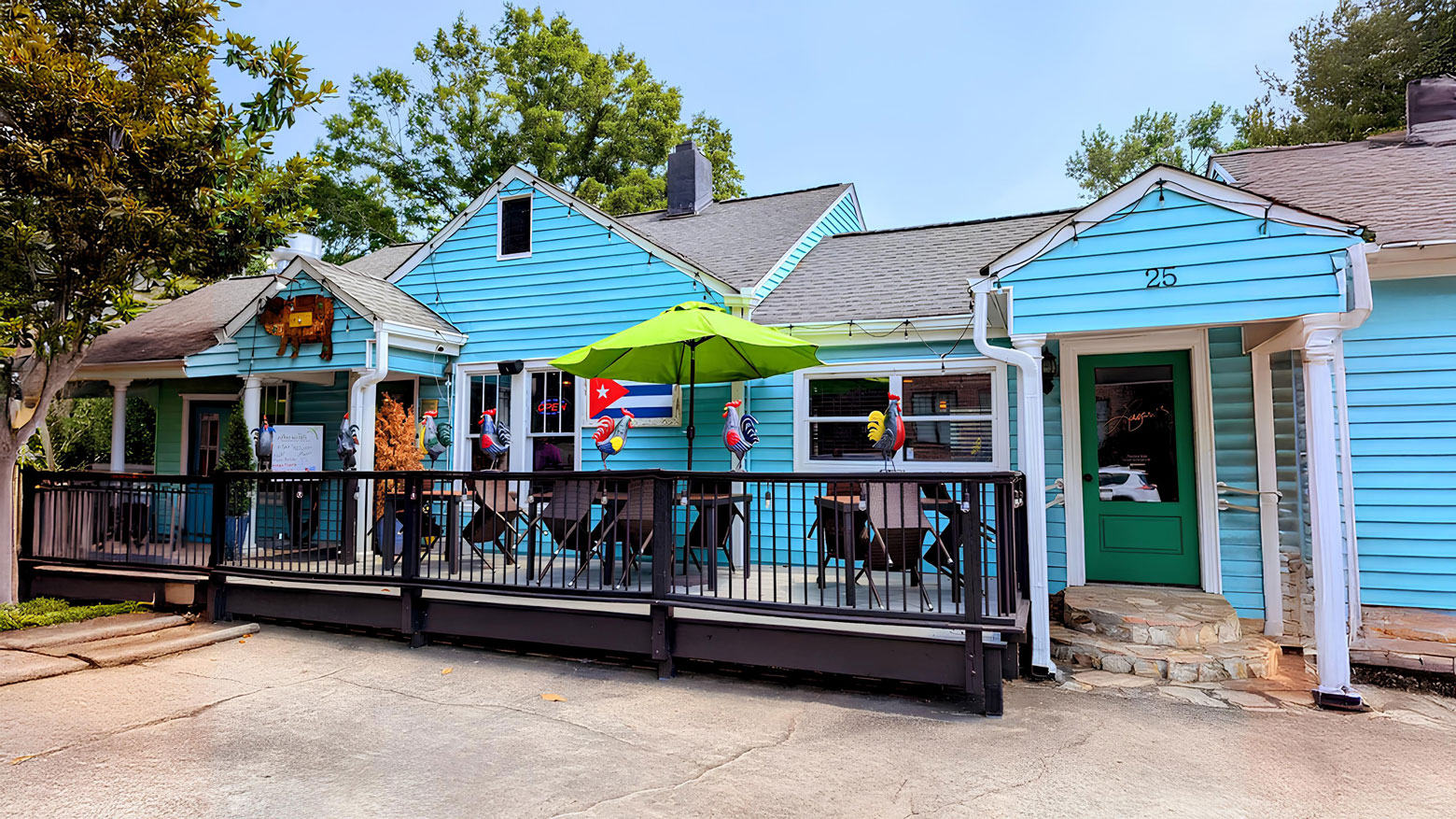 Blue building with a patio featuring colorful rooster statues and a green umbrella. Bright and inviting atmosphere with trees in the background.