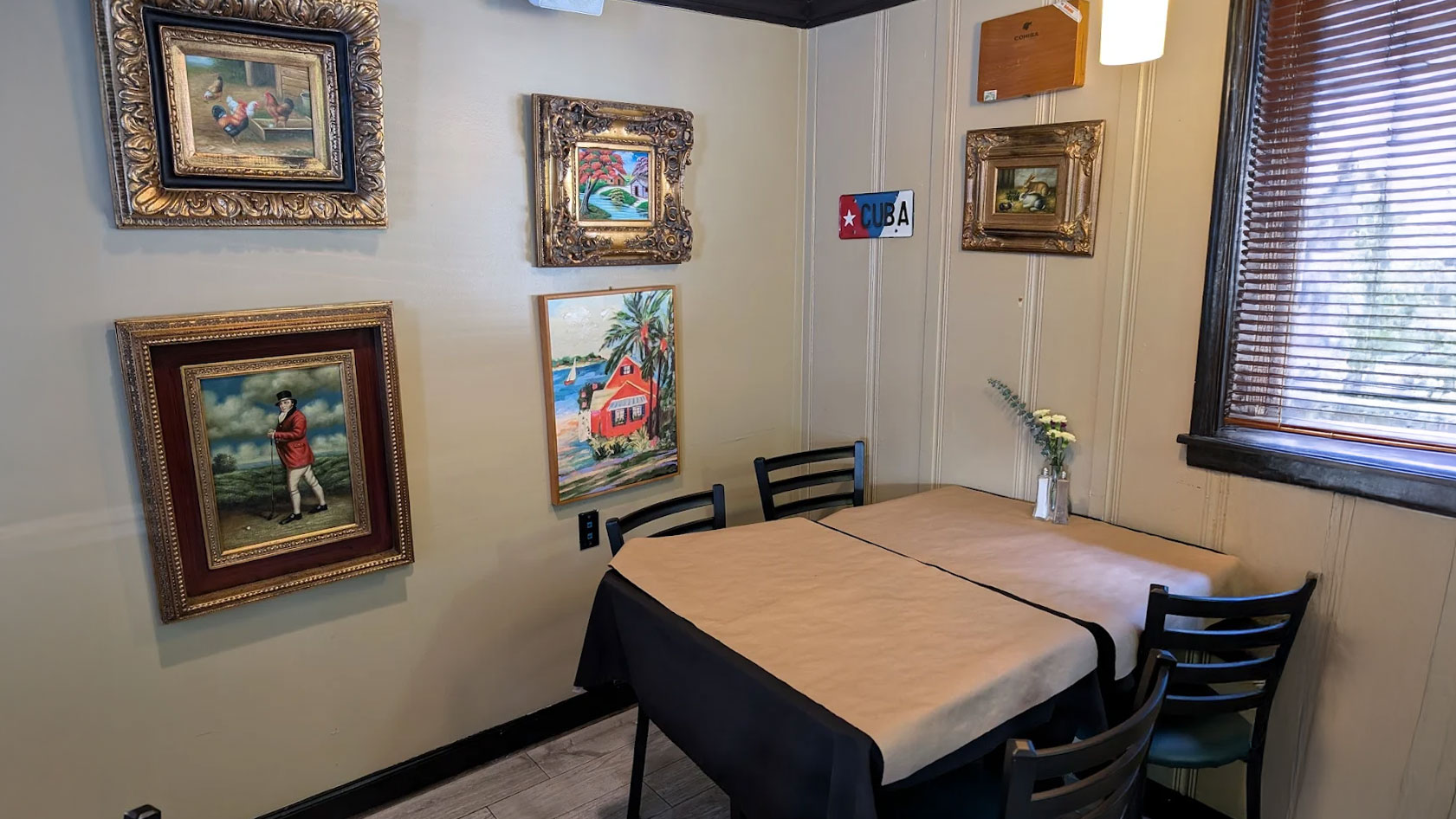 Cozy restaurant corner with two tables set for four, framed paintings of colorful landscapes and historical scenes on cream walls, a vase with flowers, and a “Cuba” sign. Warm and inviting atmosphere.