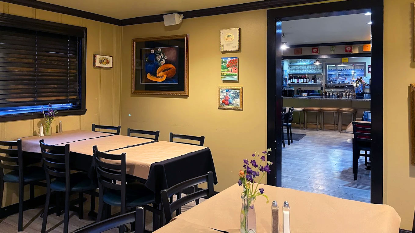 Cozy restaurant interior with wooden tables, soft lighting, and flower vases. Art decorates the walls. A bar is visible through a doorway.