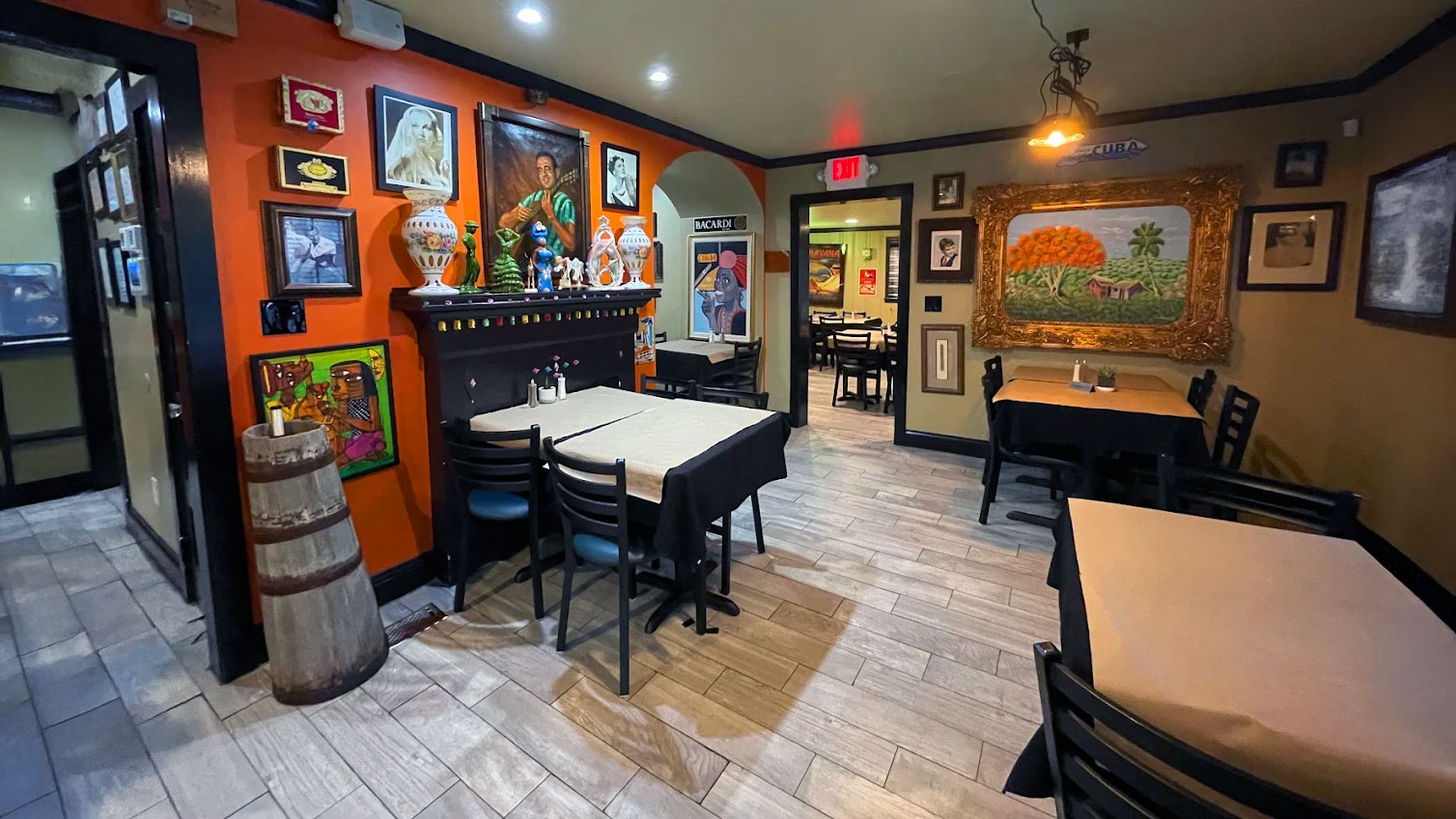 Cozy restaurant interior with orange and beige walls adorned with colorful artwork and framed paintings. Tables have black chairs and light-colored tablecloths. Warm, inviting atmosphere.