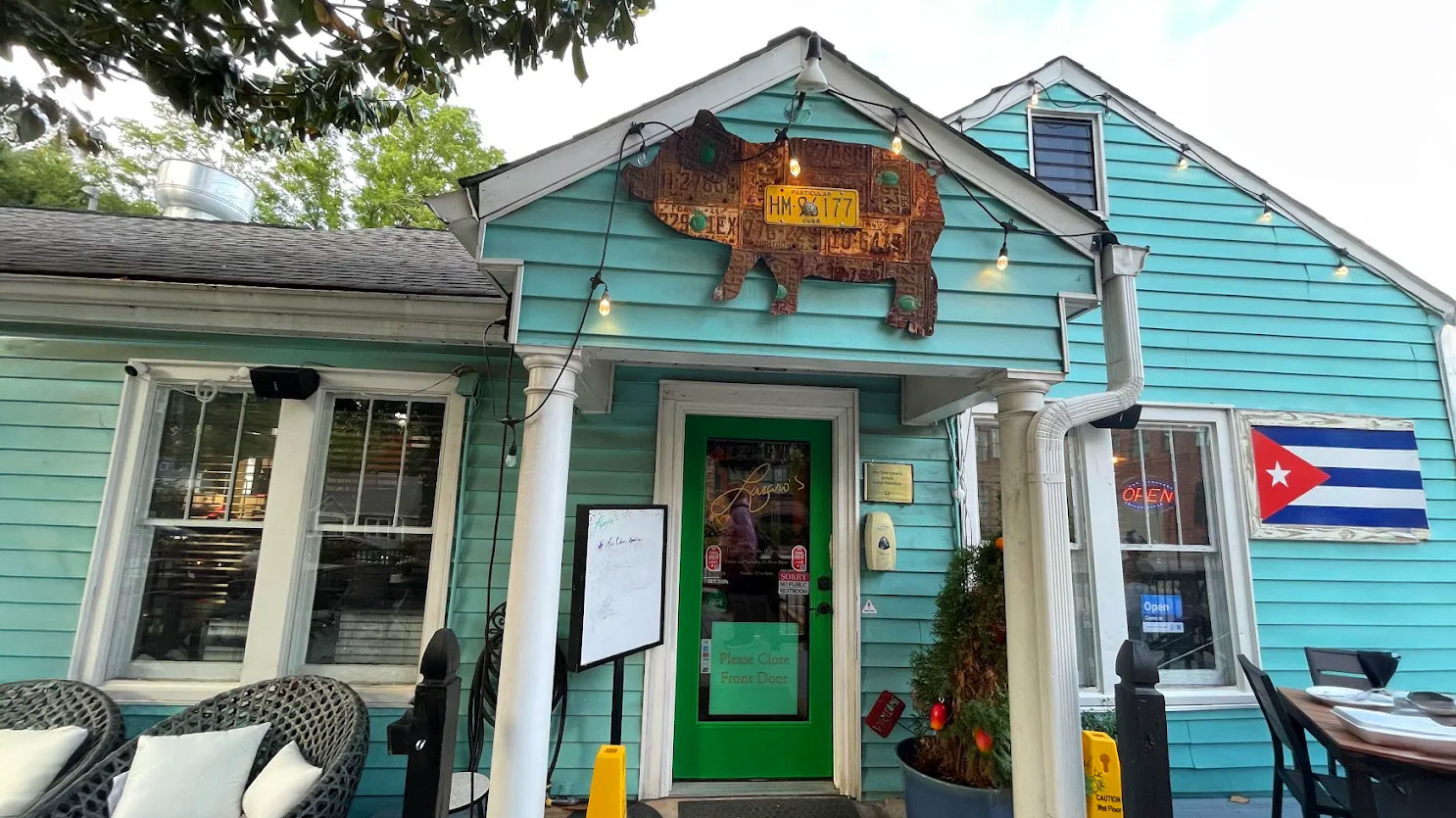 Turquoise storefront with a green door, pig-shaped sign, and a Cuban flag. Cozy outdoor seating, casual dining atmosphere.