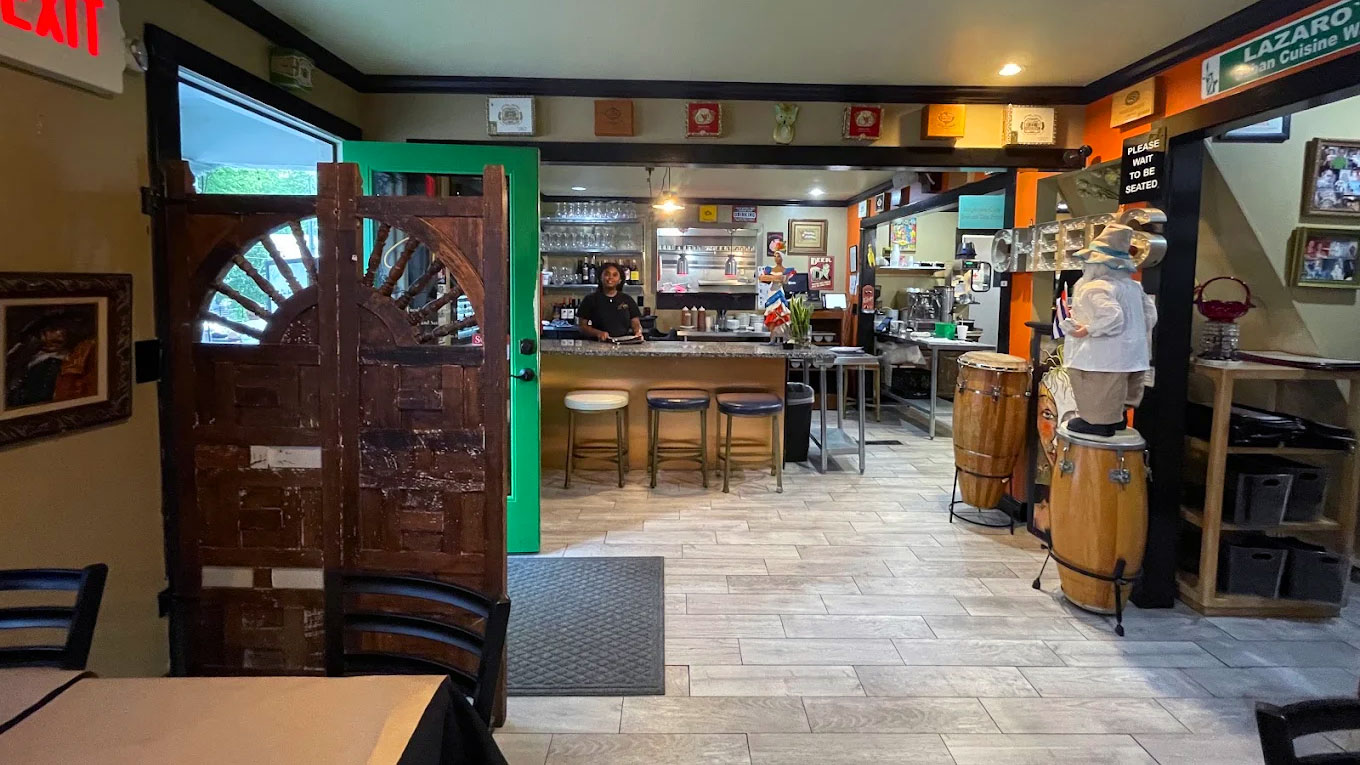A cozy restaurant interior with wooden doors, a waitress smiling behind a counter with stools. Warm lighting, conga drums, and decor create a welcoming atmosphere.