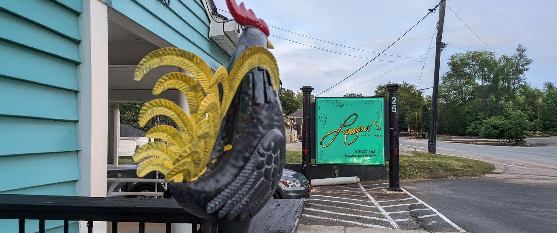 A colorful rooster statue with yellow tail feathers is in the foreground. In the background, a turquoise sign reads "Lazaro's Cuban Cuisine" near a parking lot.