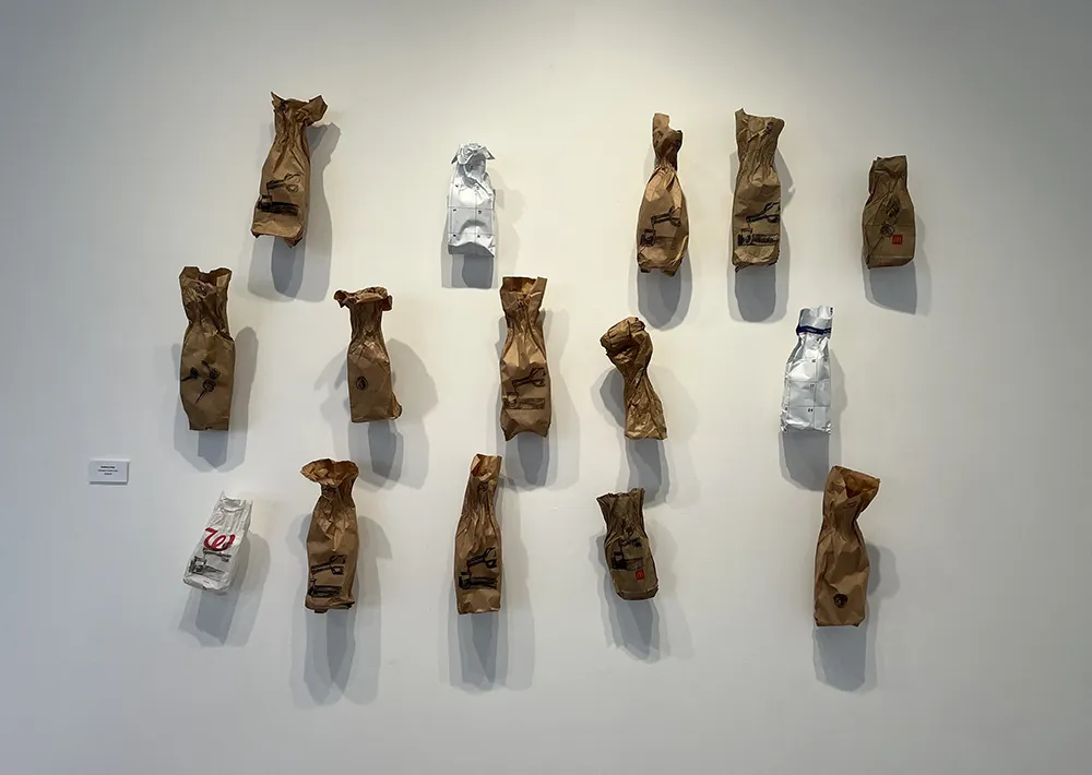 Wall installation by Robb Quisling displaying rows of brown paper and white bags pinned like specimens, each subtly crumpled and marked. The stark arrangement evokes evidence, habit, and collective memory tied to addiction.