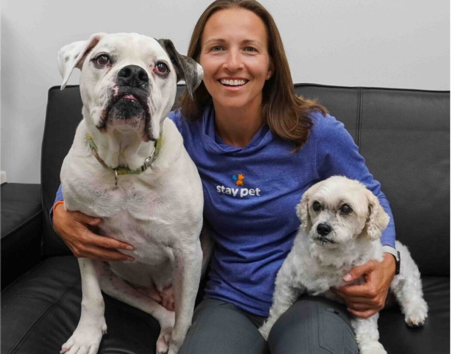 Smiling woman in a blue stay pet shirt sitting on a black couch with a large white and black dog on her left and a small white dog on her right.